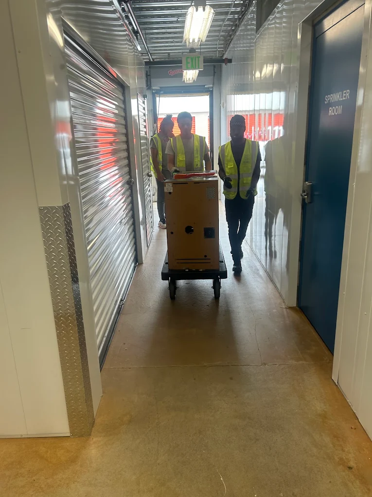 Three movers in hi-vis vests rolling a heavy cabinet down a storage facility corridor