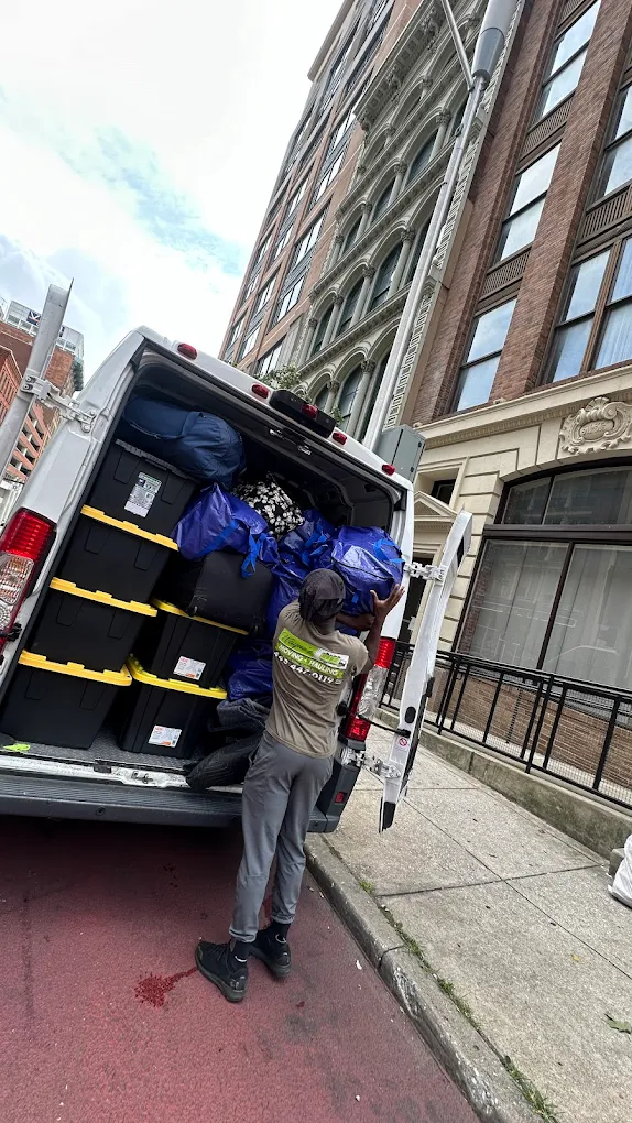 Mover loading blue-wrapped bags and black totes into a van on a downtown Baltimore street