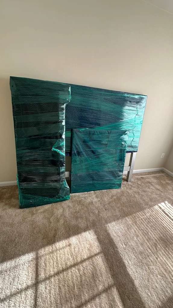 Flat furniture panels and a bed frame sealed in green stretch wrap leaning against a wall